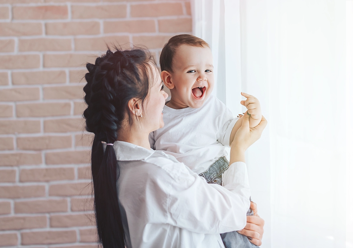 A joyful mother holding her laughing baby near a window.